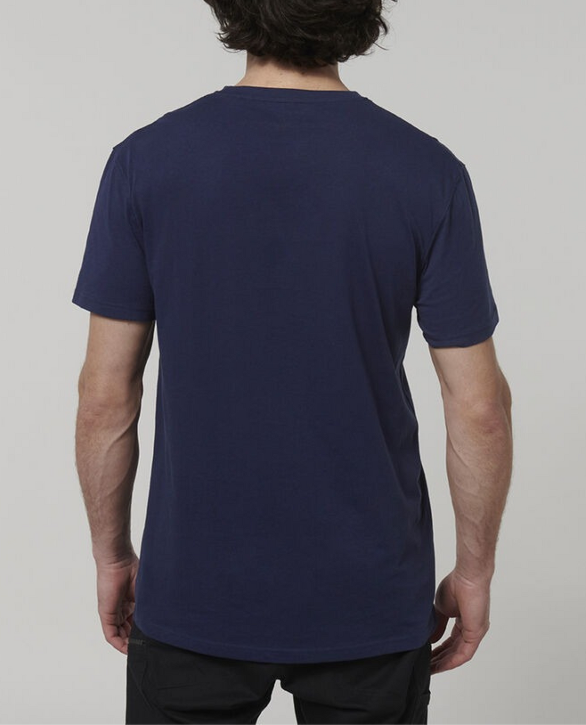 A person with short, dark hair is standing with their back to the camera, wearing a plain navy blue Hard Yakka Core Tee and black pants. The tee is made from 100% cotton. The background is a solid light grey.