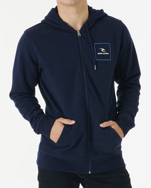 Wearing a Rip Curl Corp Icon Zip Thru hoodie in navy blue, featuring a soft fleece lining and logo patch on the chest, a person stands with hands in pockets. The hoodie showcases a white "RIP CURL" print. Black pants complete the look against a plain white background.