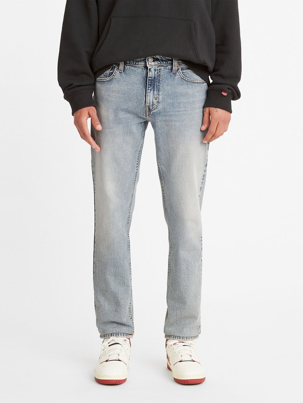 A person stands against a white background wearing a black hoodie, light blue Levis 511 Slim Jean with a slightly faded look, exuding effortless style, and white sneakers with red accents. Their hands are relaxed by their sides.