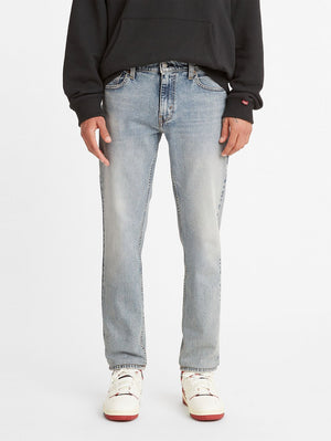 A person stands against a white background wearing a black hoodie, light blue Levis 511 Slim Jean with a slightly faded look, exuding effortless style, and white sneakers with red accents. Their hands are relaxed by their sides.