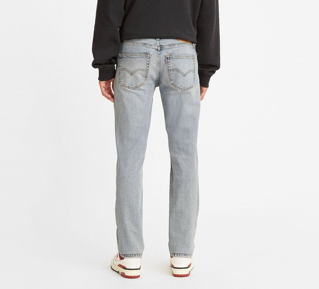 Rear view of a person in Levi's 511 Slim jeans, light blue with straight legs, two visible back pockets. They're wearing a black sweatshirt and white sneakers with red accents against an unadorned white backdrop.