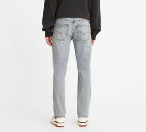 Rear view of a person in Levi's 511 Slim jeans, light blue with straight legs, two visible back pockets. They're wearing a black sweatshirt and white sneakers with red accents against an unadorned white backdrop.