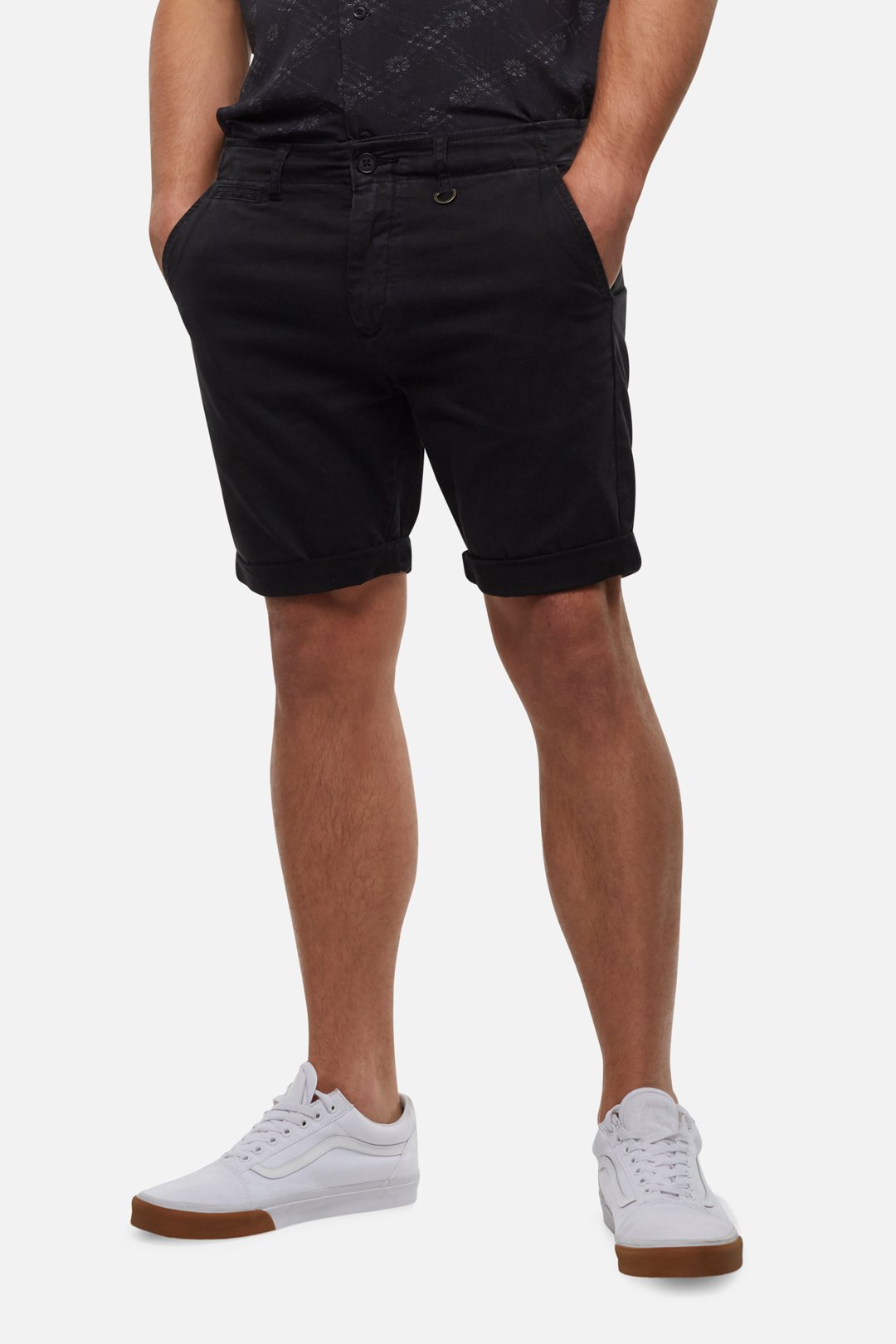 A person is standing against a plain background, wearing Industrie The Rinse Short in black, a dark-patterned shirt, and white sneakers. The person has their hands comfortably in their pockets. Only the lower half of their body is visible.