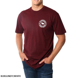 A person wearing the Ringers Western Signature Bull Original Fit T-Shirt in burgundy, made of 100% cotton, featuring a white circular logo with a bull and the text "Ringers Western" on the left chest. One hand is in their pocket while the other hangs by their side. Text reads "BURGUNDY/WHITE.