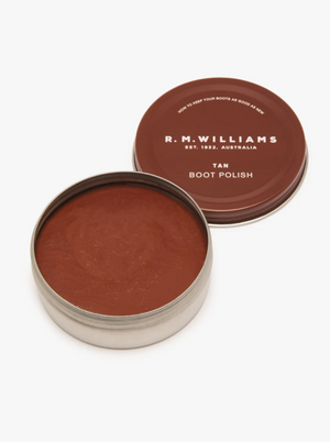 A round, open tin of RM Williams Stockmans Boot Polish is placed with its lid beside it. The lid is brown with white text that reads, "R.M.WILLIAMS, Est. 1932, Australia, Tan Boot Polish." The alcohol-free formula inside the tin helps restore color and shine to your boots seamlessly.