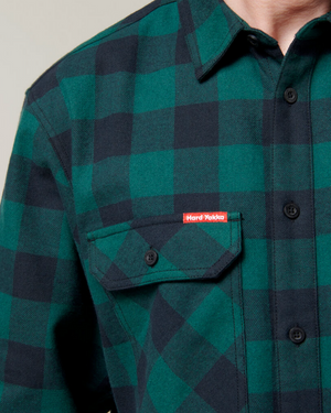 Close-up of a person wearing the Hard Yakka Check Flannelette Shirt, made from durable cotton twill in green and black checks, featuring black buttons and a front pocket with a small red "Hard Yakka" label.