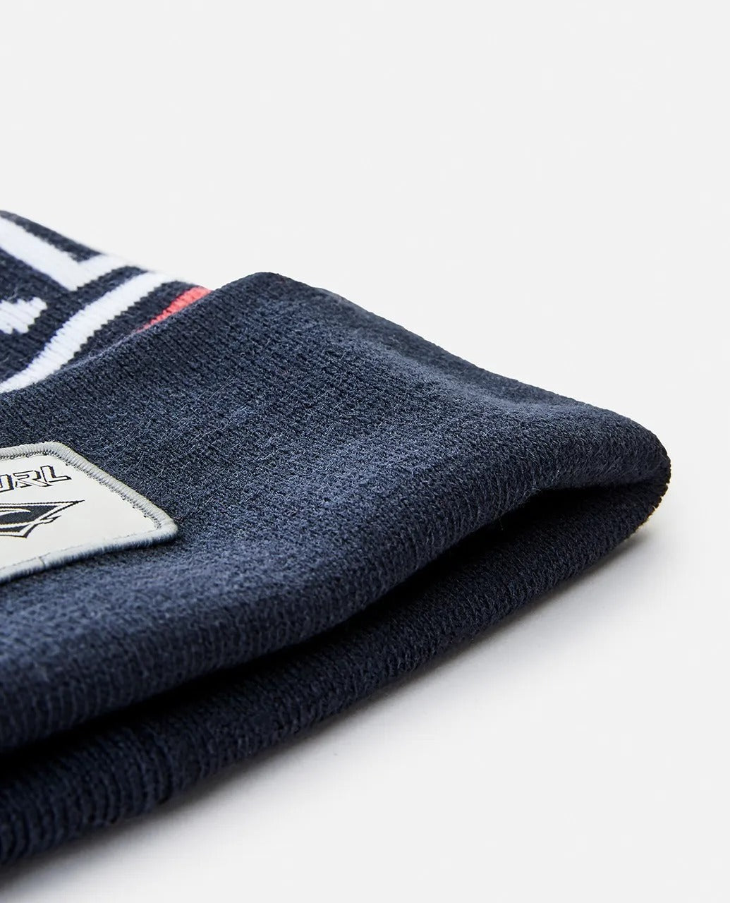 A close-up of a navy blue Rip Curl Fade Out Tall Beanie featuring a white and red pattern on its edge. A rectangular fabric label with the logo is stitched onto the side. The background is plain and light-colored.