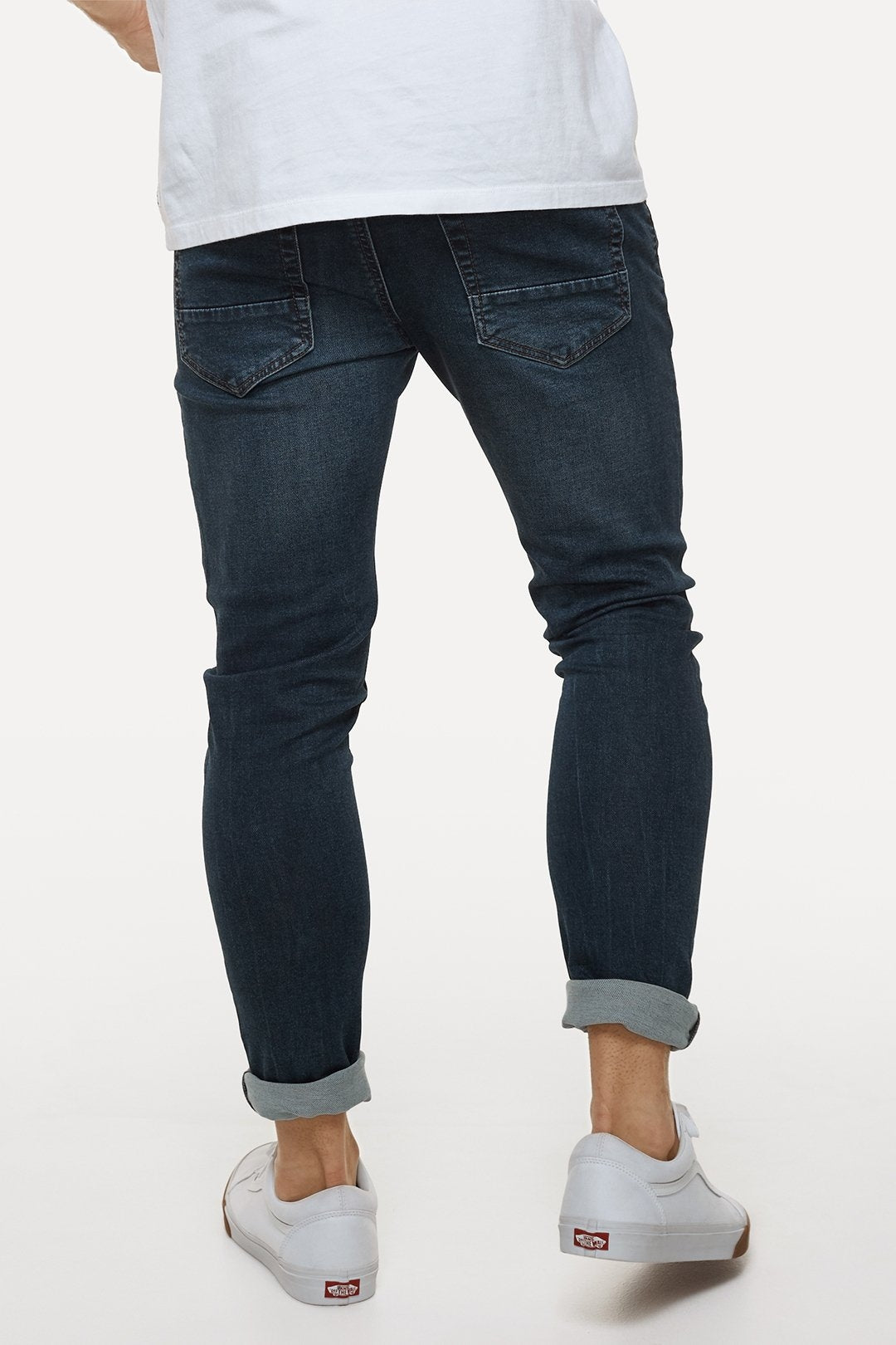 A person wearing dark blue Industrie The Denim Drifter NC Pant and a white T-shirt is seen from the back. The jeans are rolled up at the ankles, revealing white sneakers with red circular logos. The style complements the plain white background effortlessly.