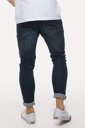 A person wearing dark blue Industrie The Denim Drifter NC Pant and a white T-shirt is seen from the back. The jeans are rolled up at the ankles, revealing white sneakers with red circular logos. The style complements the plain white background effortlessly.