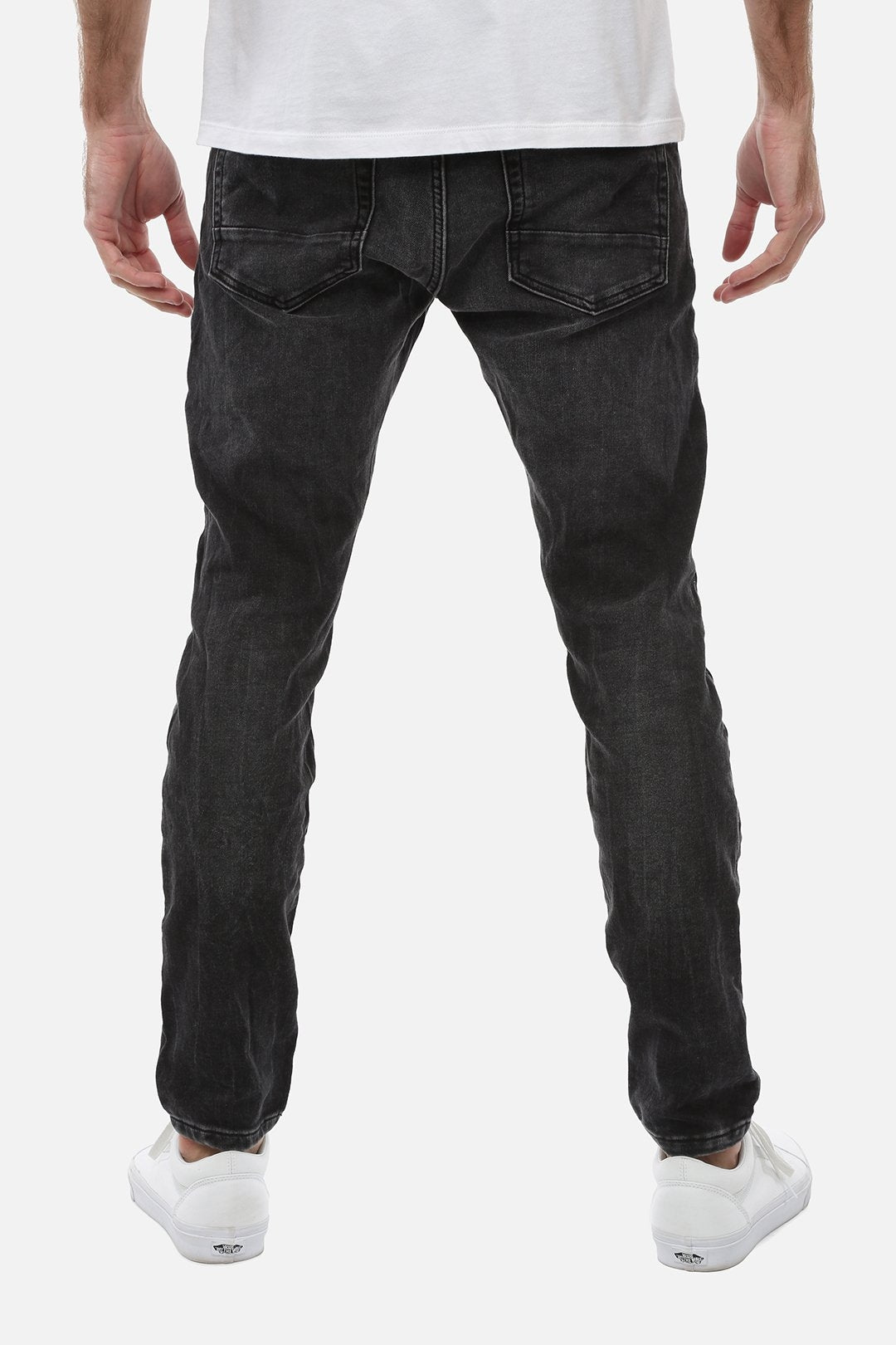 Back view of a person wearing the Industrie The Denim Drifter NC Pant in black denim, complemented by white sneakers. The person is also wearing a white shirt, partially visible at the top of the image. Only the lower half of the person's body is shown against a plain white background.