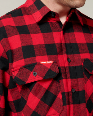 Close-up of a person wearing a red and black plaid Hard Yakka Check Flannelette Shirt made from durable cotton twill, featuring buttoned flap pockets. The small "Hard Yakka" label is visible on one pocket, emphasizing the shirt's pattern, texture, and UPF 50+ protection.