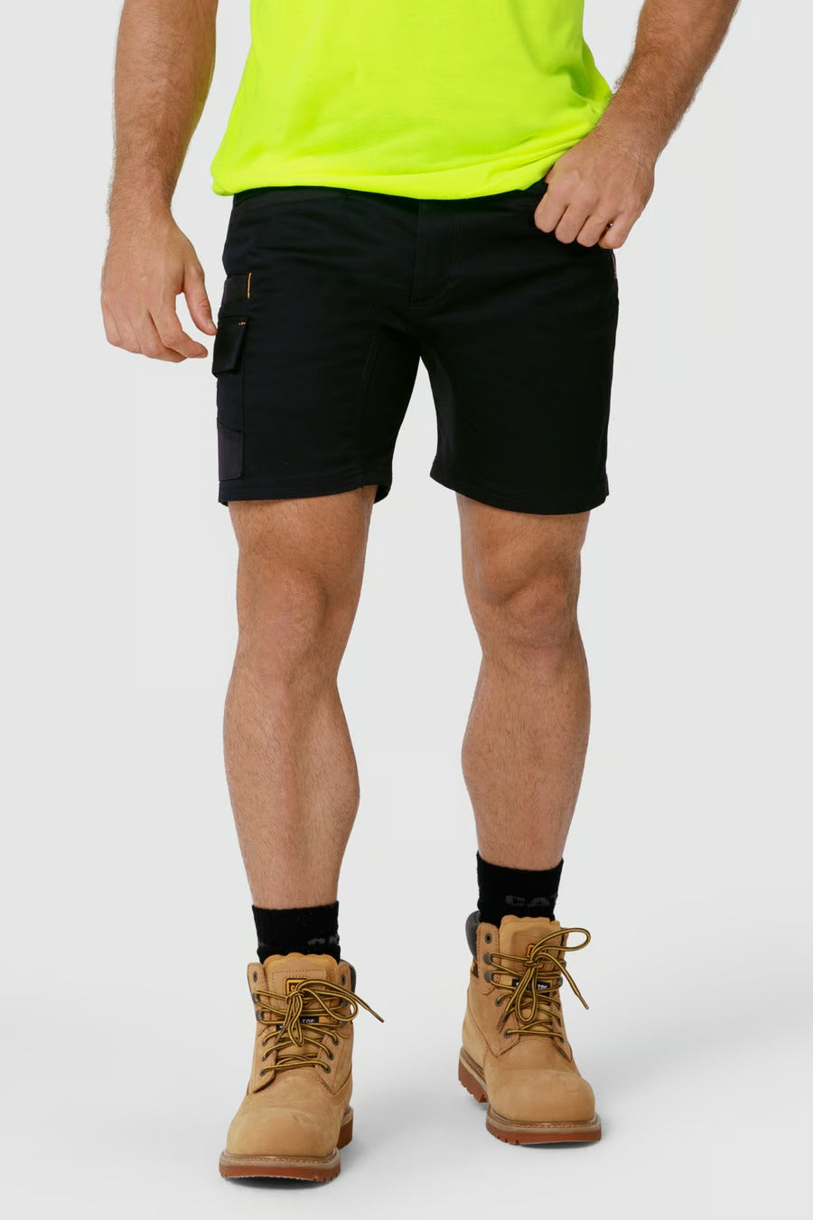 A person wearing a bright neon green shirt, CAT Elite Operator Shorts in black with utility pockets, black socks, and tan work boots stands against a plain light gray background. The upper body is not visible in the image.