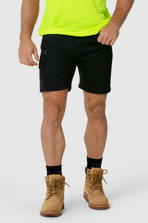 A person wearing a bright neon green shirt, CAT Elite Operator Shorts in black with utility pockets, black socks, and tan work boots stands against a plain light gray background. The upper body is not visible in the image.