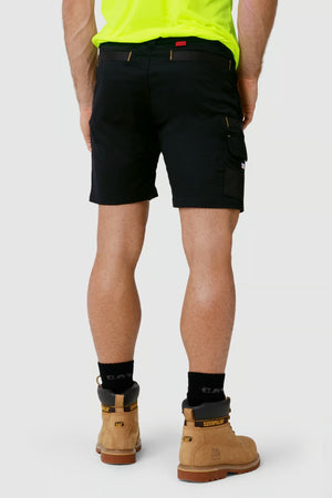 A person is standing and facing away, wearing CAT Elite Operator Short in black with multiple utility pockets, a neon green shirt, black socks, and tan work boots. The background is plain gray.