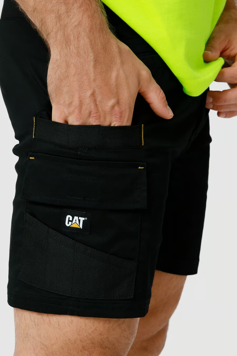 Close-up of a person wearing black CAT Elite Operator Shorts, which feature multiple utility pockets and a "CAT" logo patch on one of the pockets. The individual is also sporting a bright yellow shirt, tucked into the shorts with elastic inserts. Only the midsection and upper thighs are visible.