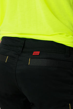 A close-up image features a person wearing the CAT Elite Operator Short in black, showcasing its utility pockets. The individual is also sporting a bright yellow top. A small red tag with text is visible at the back of the waistband on the shorts, though it is not fully readable in the image.