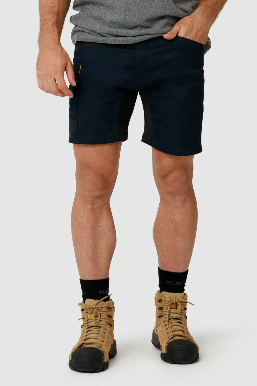 A person wears a gray t-shirt and dark CAT Elite Operator Shorts with utility pockets, complemented by black socks and beige hiking boots. Their left hand is tucked into a pocket while the right hand rests by their side. The background is plain white.