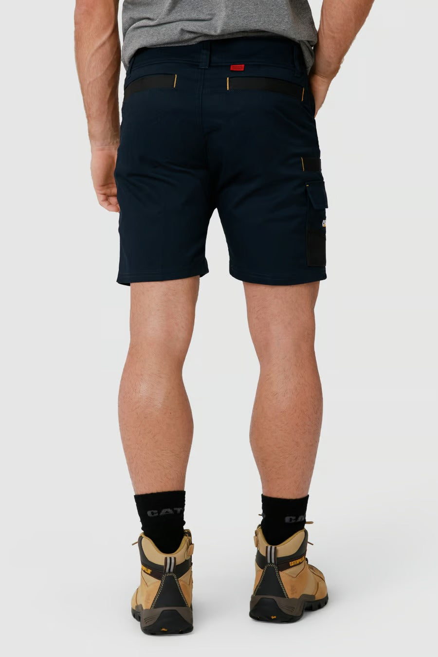 A person is standing with their back to the camera, wearing the CAT Elite Operator Short in navy blue, which features utility pockets and a small red tab on the waistband. Paired with a gray T-shirt, black socks, and tan hiking boots, they stand against a plain white background.
