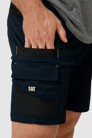 A person wearing a pair of dark blue CAT Elite Operator Shorts is shown. The shorts feature multiple utility pockets, including a velcro flap pocket with the CAT logo and an additional smaller pocket above it. The individual, dressed in a grey shirt, is placing their hand into one of the pockets.