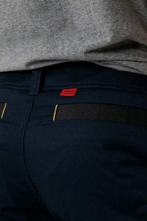 A close-up image showcasing the dark blue CAT Elite Operator Short, featuring yellow stitching on the utility pocket and a small red tag above it. The person is also wearing a gray shirt.