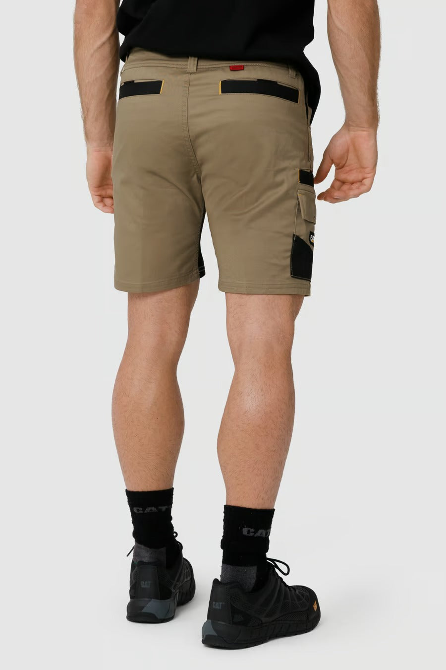 A person is wearing a black T-shirt, CAT Elite Operator Shorts in tan with elastic inserts and black and yellow accents on the utility pockets, black socks, and black shoes. They are shown from the back, displaying the details of the shorts and footwear. The background is plain white.