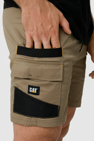 A person wearing CAT Elite Operator Shorts with multiple utility pockets is placing their hand into one of the pockets. The tan shorts, boasting elastic inserts for added comfort, feature a "CAT" logo on a black and yellow label. The background is plain and light-colored.