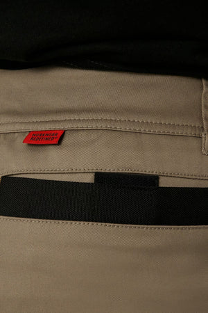 Close-up image of the back pocket of a pair of CAT Elite Operator Shorts in khaki stretch fabric, featuring black details. A small red tag near the pocket seam displays "WISCONSIN LIGHTHOUSE." The waistband, elastic inserts, and belt loops are also visible.