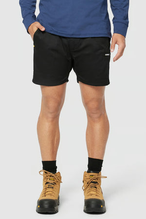 A person modeling the CAT Short Haul Short, featuring a small white logo on the left leg, paired with a blue long-sleeve shirt. The individual is also wearing black socks and beige hiking boots, standing against a plain light background.