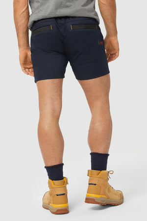 A person wearing a grey t-shirt, CAT Short Haul Shorts in navy blue with an elastic waist and black zippered pockets, and beige lace-up work boots with blue socks. The view is from the back, showing the person's legs and feet against a plain white background.