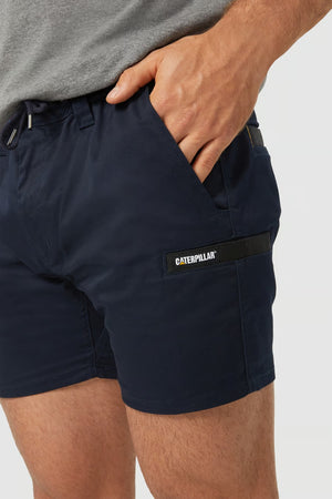 A person is shown from the waist down wearing a pair of navy blue CAT Short Haul Shorts. The shorts feature an elastic waistband, convenient side pockets, and a cargo pocket with a Caterpillar logo patch on the left thigh. The person has a hand casually placed in one of the pockets.