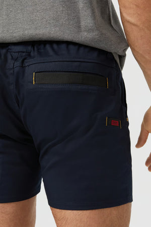 A person wearing the CAT Short Haul Short in navy blue and a grey shirt is shown. A close-up image features the back cargo pocket of the shorts, which has a flap secured by a black strip and yellow stitching. The fabric tag beside the pocket is visible with red and black details.