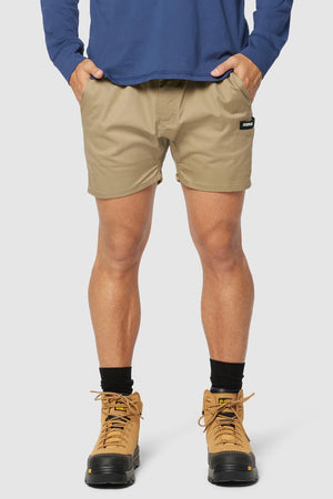 A person wearing a blue long-sleeved shirt, CAT Short Haul Short in beige with cargo pockets and hands in pockets, black socks, and tan lace-up boots. The background is plain white.