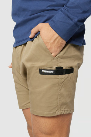 A person wearing the CAT Short Haul Shorts, which are khaki-colored with a cargo pocket and an elastic waist. Their right hand is in the pocket that has a "Caterpillar" brand label on it. They are also dressed in a blue long-sleeve shirt.