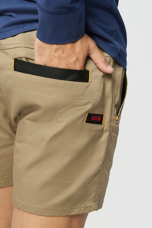 A person wearing a blue long-sleeve shirt and CAT Short Haul Shorts, which are beige with yellow stitching. The person's hand is placed in the back pocket of the shorts, which feature an elastic waist. There is a small black and red tag above the pocket.