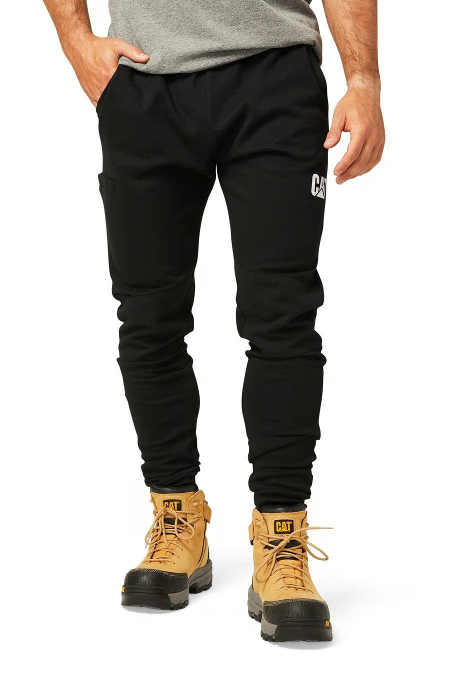 A person is shown from the shoulders down wearing CAT Track Pant, featuring a small logo on the left thigh. They are also sporting a gray shirt and tan work boots with black toes. The background is white.