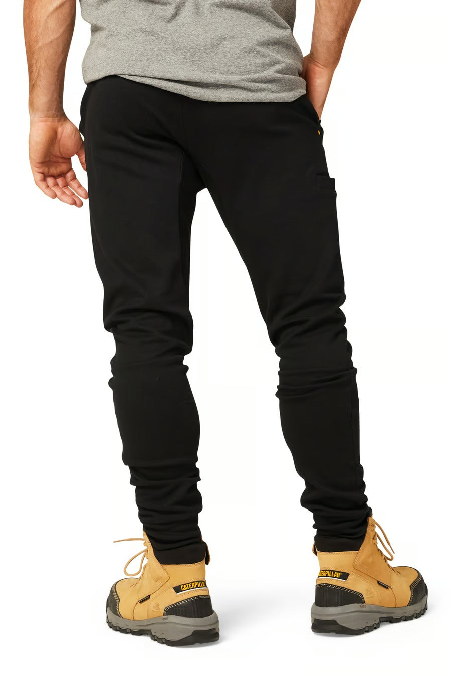 A person wearing black CAT Track Pants, a gray shirt, and tan work boots with yellow laces and a black and yellow label on the back of the boots. The image shows the person from behind, highlighting the back of the pants and the boots.