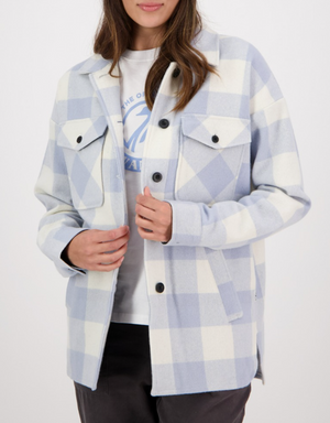 A woman models the Swanndri Anchorage Wool Shirt Jacket in light blue and white plaid over a white graphic tee and dark pants, her hands near the jacket's buttons. Her face is partly out of frame.