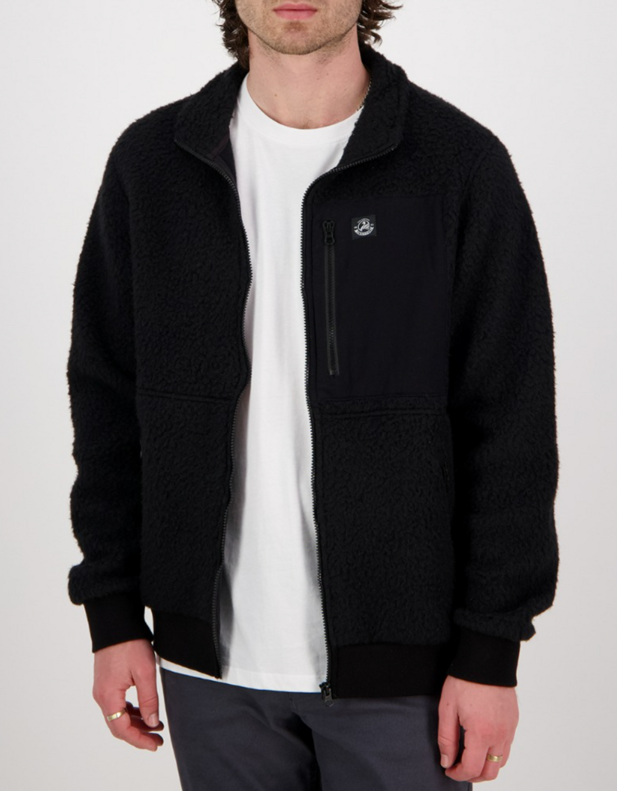 Someone is wearing a Swanndri Churchill Wool Fleece Jacket in black over a white T-shirt and gray pants. The cozy jacket has a small chest pocket and a subtle emblem, set against a plain white background.