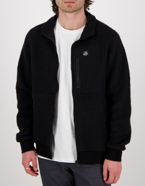 Someone is wearing a Swanndri Churchill Wool Fleece Jacket in black over a white T-shirt and gray pants. The cozy jacket has a small chest pocket and a subtle emblem, set against a plain white background.