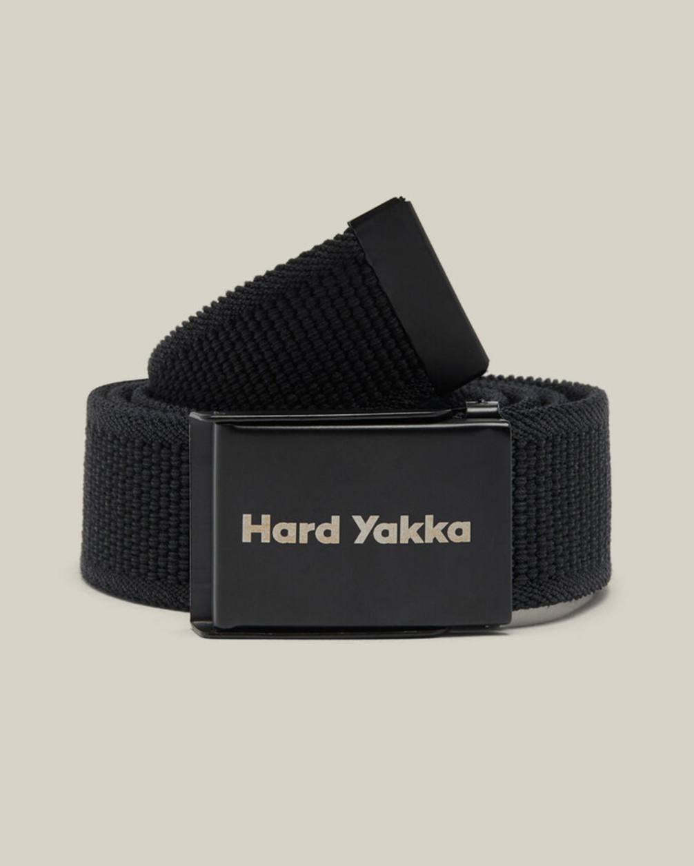 A black Hard Yakka Stretch Webbing Belt is coiled neatly, showcasing its rectangular metal buckle that doubles as a bottle opener and reads "Hard Yakka" in white text. The plain beige background highlights the belt.