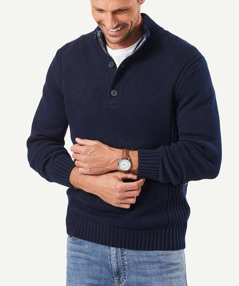 A person is smiling and looking down, dressed comfortably in the Gazman Chunky Button Neck Knit over a white t-shirt and checked collared shirt. They are also wearing light blue jeans and a brown wristwatch on their left wrist.
