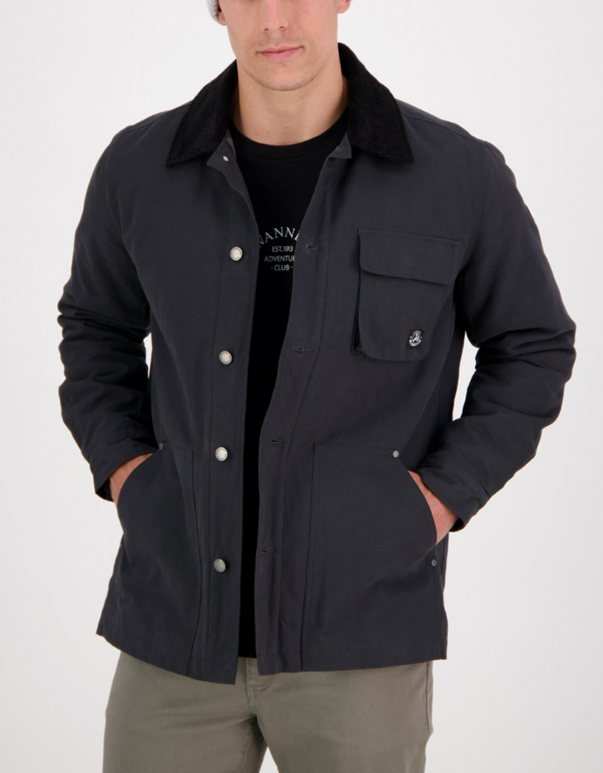 A person in a Swanndri Barrytown v2 Canvas Work Jacket with a button-up front, dark blue canvas, black collar, quilted lining, and an anchor emblem on the chest pocket. They're wearing a beanie, gray pants, and a black T-shirt with their hands in the jacket pockets.