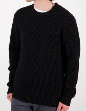 A person wears the Swanndri Fistral Waffle Knit black sweater over a white shirt with dark pants. The photo is cropped at the neck and mid-thigh, set against a plain white background.