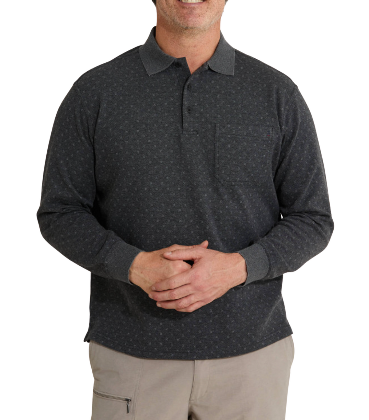 A person with short hair is wearing a Breakaway Lander Micro Prima Polo, a long-sleeved dark gray shirt featuring a subtle pattern and chest pocket. Their hands are clasped in front, paired with light khaki pants, and their face remains unseen.