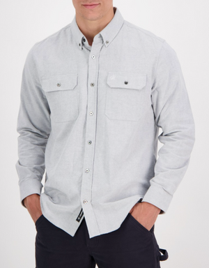 A person sports a Swanndri Marwood Shirt in light gray, a stylish classic-fit button-up made of 100% cotton. It features two chest pockets, and their hands rest casually in the pockets of their dark pants against a plain white background.