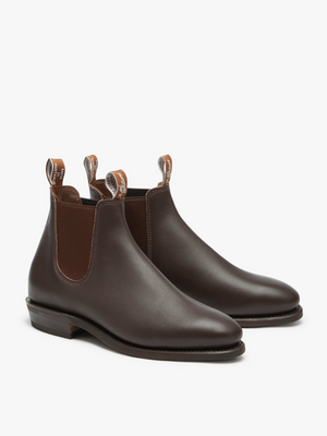 The RM Williams Adelaide Rubber Sole Boots are brown leather with elastic side panels and pull tabs at the back and front, offering a smart-casual appeal. They feature a sleek, polished finish and a low heel, showcased against a pristine white background.