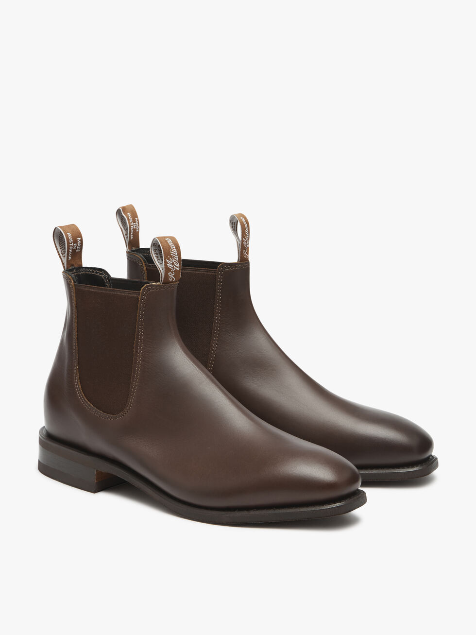This is the RM Williams Comfort Craftsman Boot, a pair of handcrafted brown leather Chelsea boots featuring black elastic side panels and pull tabs at both the back and front, adorned with white text. Crafted from yearling leather with a sleek, polished finish, these boots offer a low heel and a comfort innersole. The image showcases the boots against a plain white background.