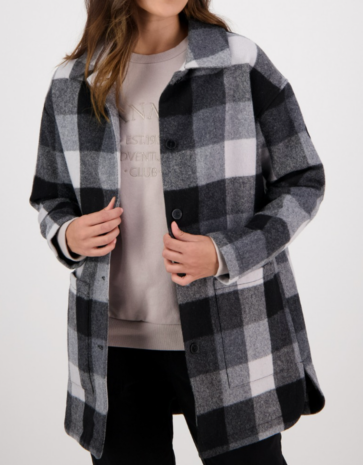 A person is adjusting a Swanndri Becroft Wool Coat, a stylish, comfortable plaid in black, white, and gray over a beige sweatshirt. This merino wool blend comes with a collar and buttons down the front. Their face is hidden against a plain light background.