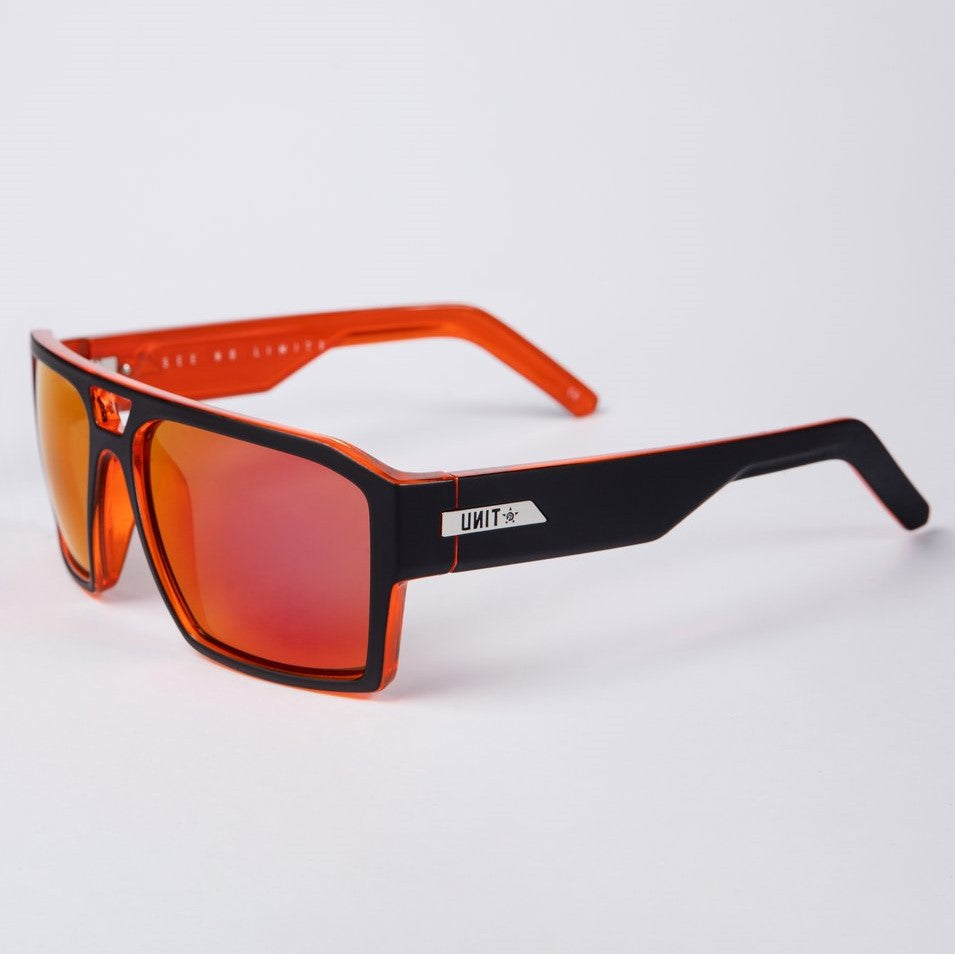 The UNIT Vault Polarised Eyewear features rectangular frames with red-tinted lenses. The front of the frames are black, transitioning to orange along the sides, and provide excellent protection against sun glare. A silver brand logo displaying "UNIT" and a star adorns the temples. The background is plain white.