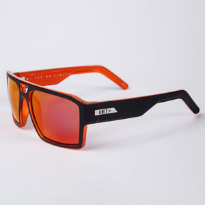 The UNIT Vault Polarised Eyewear features rectangular frames with red-tinted lenses. The front of the frames are black, transitioning to orange along the sides, and provide excellent protection against sun glare. A silver brand logo displaying "UNIT" and a star adorns the temples. The background is plain white.